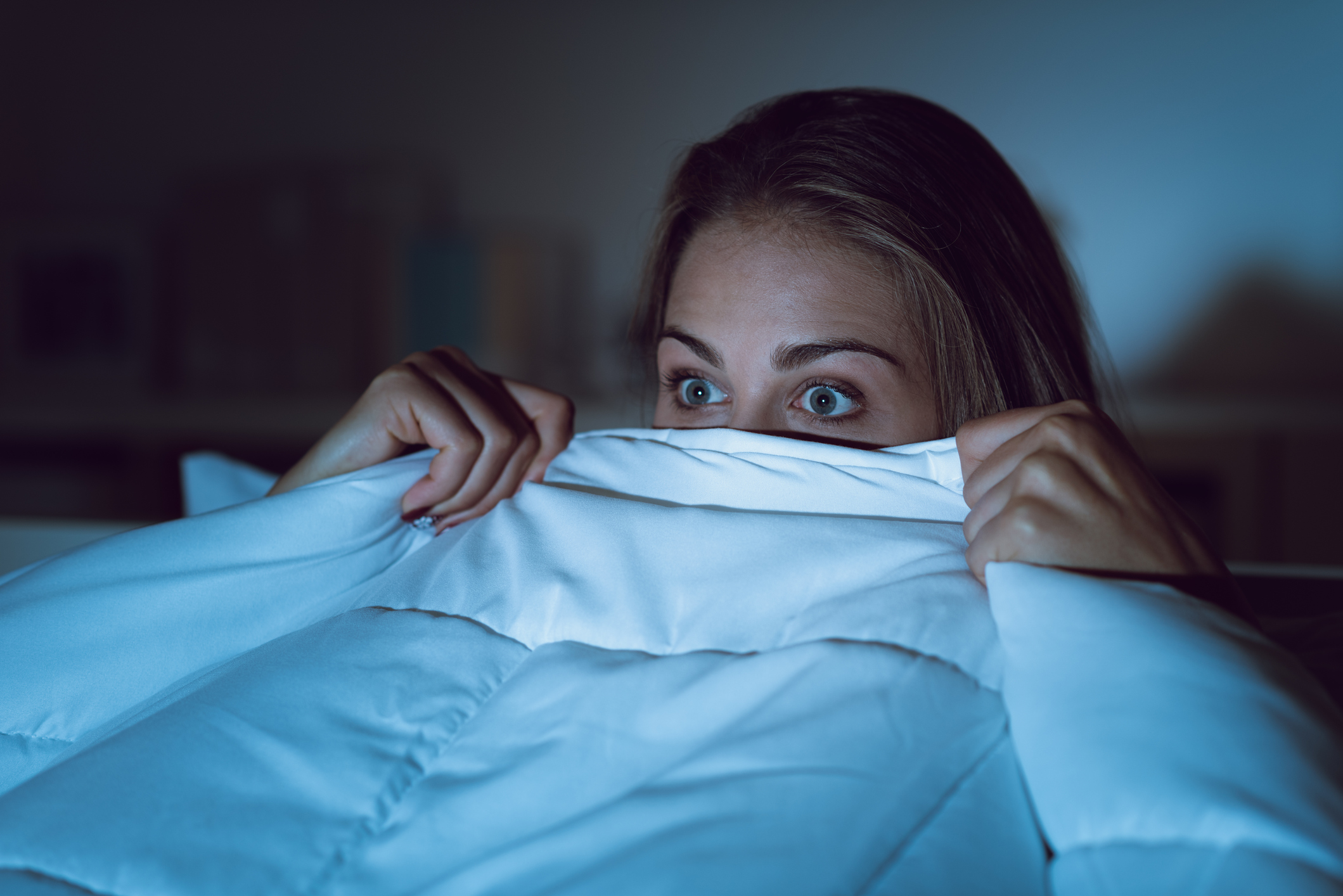 Woman watching a scary horror movie on tv late at night, she is frightened and hiding under the blanket | FintechZoom